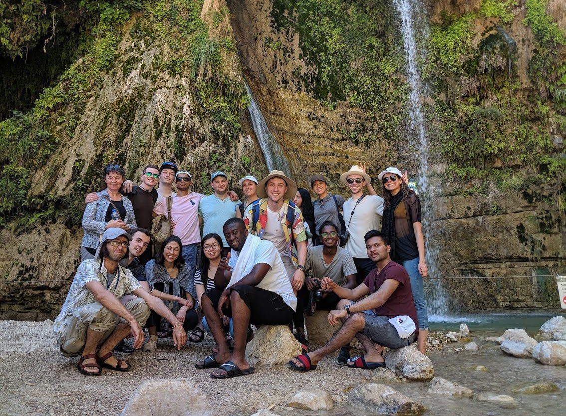 a diverse group of smiling people stand and sit on the rocks at the base of a waterfall. All of the people smile for photo.